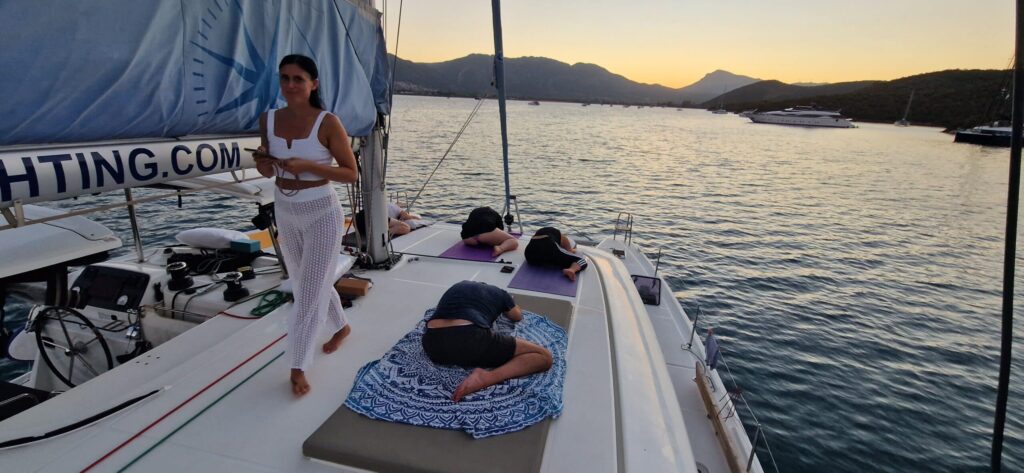 Eine Frau in weißem Outfit steht bei Sonnenuntergang auf dem Deck eines Segelboots und hält eine Tasse in der Hand. Zwei Personen machen Yoga-Posen auf Matten und eine dritte Person liegt auf einem Handtuch. Das Segelboot ist Teil eines Skippertrainings im Mittelmeer und schwimmt ruhig mit anderen Booten und Bergen im Hintergrund.