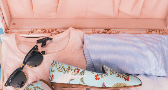 Ein rosa Koffer voller ordentlich angeordneter Gegenstände, darunter ein pastellfarbenes T-Shirt, ein gestreiftes Hemd, ein Paar Schuhe mit Blumenmuster und eine schwarze Sonnenbrille für eine Segelreise.