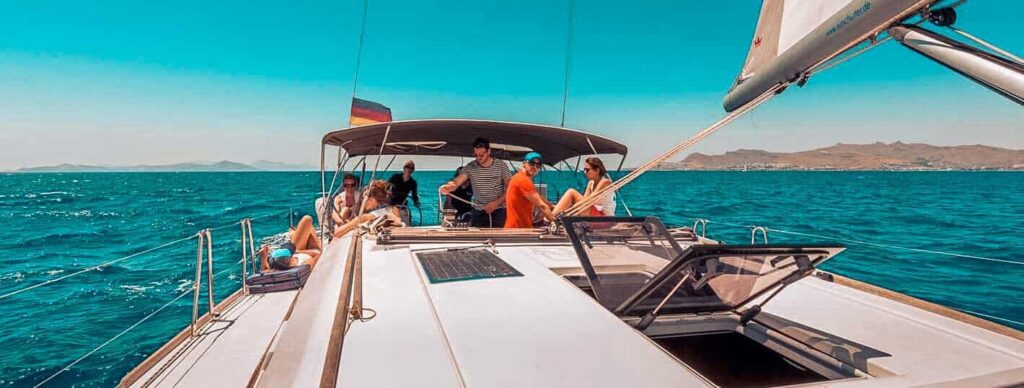 Eine Gruppe von Menschen genießt einen sonnigen Tag auf einem Segelboot und segelt über blaue Gewässer, im Hintergrund sind in der Ferne Berge zu sehen.