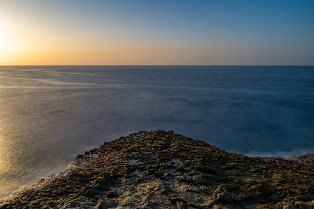 Ein felsiger Vordergrund führt zu einem weiten, ruhigen Ozean unter klarem Himmel. Die Sonne geht entweder auf oder unter und wirft einen warmen Schein auf den Horizont, der sich subtil im Wasser spiegelt. Die Szene ist heiter und weitläufig und ruft ein Gefühl der Ruhe hervor – fast wie ein automatisch gespeicherter Entwurf der Schönheit der Natur.