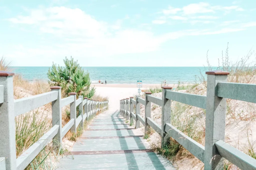 Ein Holzsteg mit Geländer führt zu einem Sandstrand und ruhigem Meer unter einem klaren Himmel, mit spärlichem Grün an den Seiten und einer Segelyacht in der Nähe des Wassers.